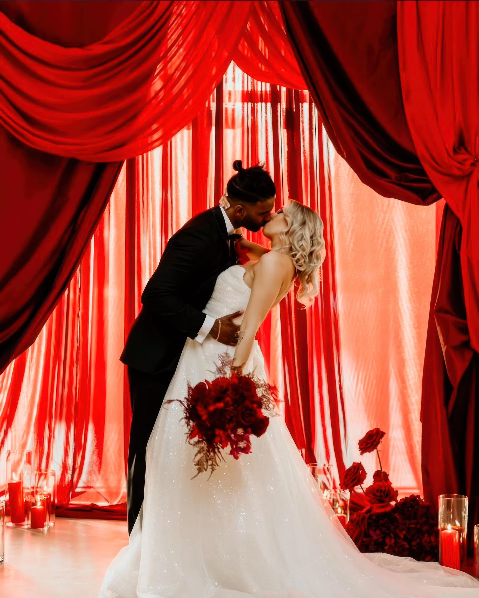 Bride and groom kissing beneath red draped wedding arch with candles and roses, wedding decor hire Worcestershire