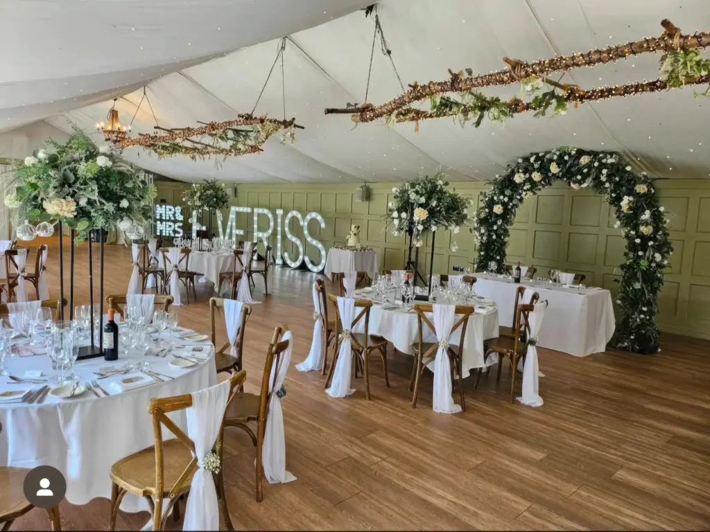 Wedding decor hire Hogarths Hotel Solihull — illuminated surname letters, floral wedding arch, tall table centrepieces with greenery, rustic ceiling decor, cross-back chairs with white sashes