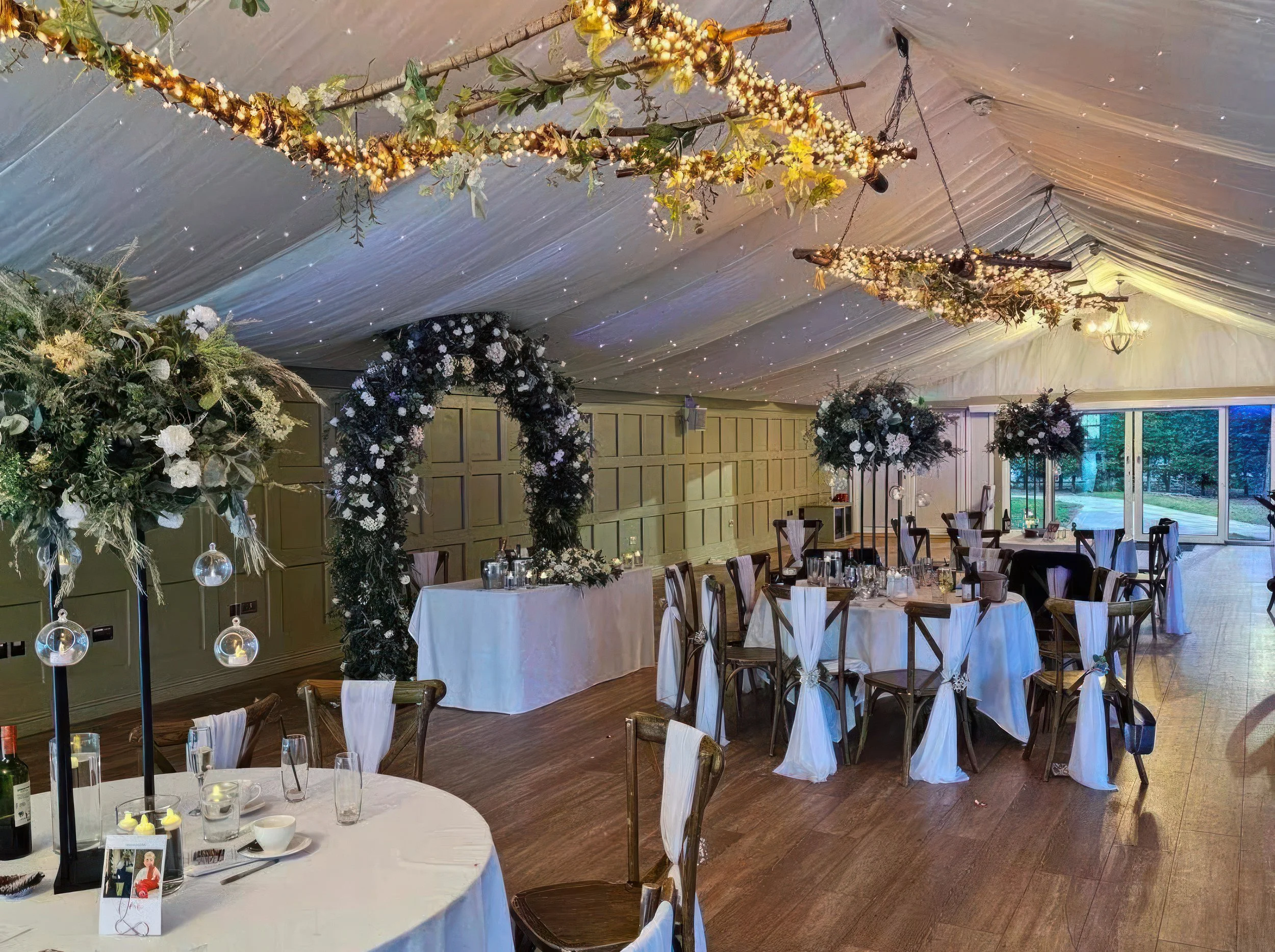 Wedding decor hire Hogarths Hotel Solihull — winter floral arch, tall table centrepieces, ceiling decor with fairy lights and foliage, cross-back chairs with white sashes