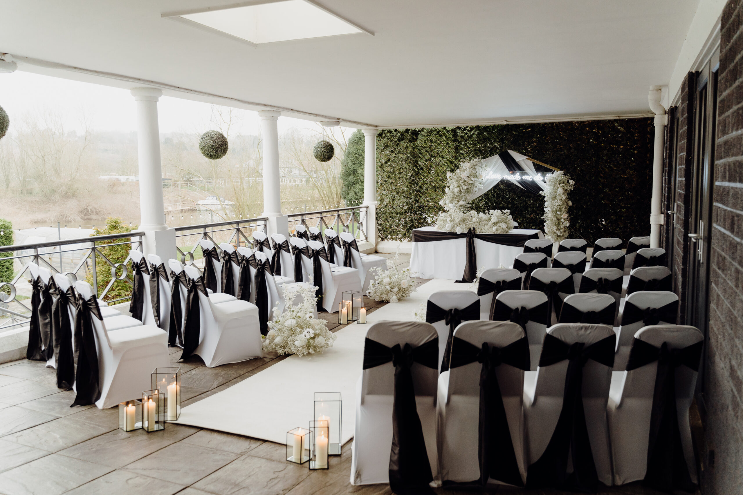 Outdoor wedding ceremony with white chair covers, black sashes, lanterns, floral arch and flower wall at Holt Fleet, wedding decor hire Worcestershire