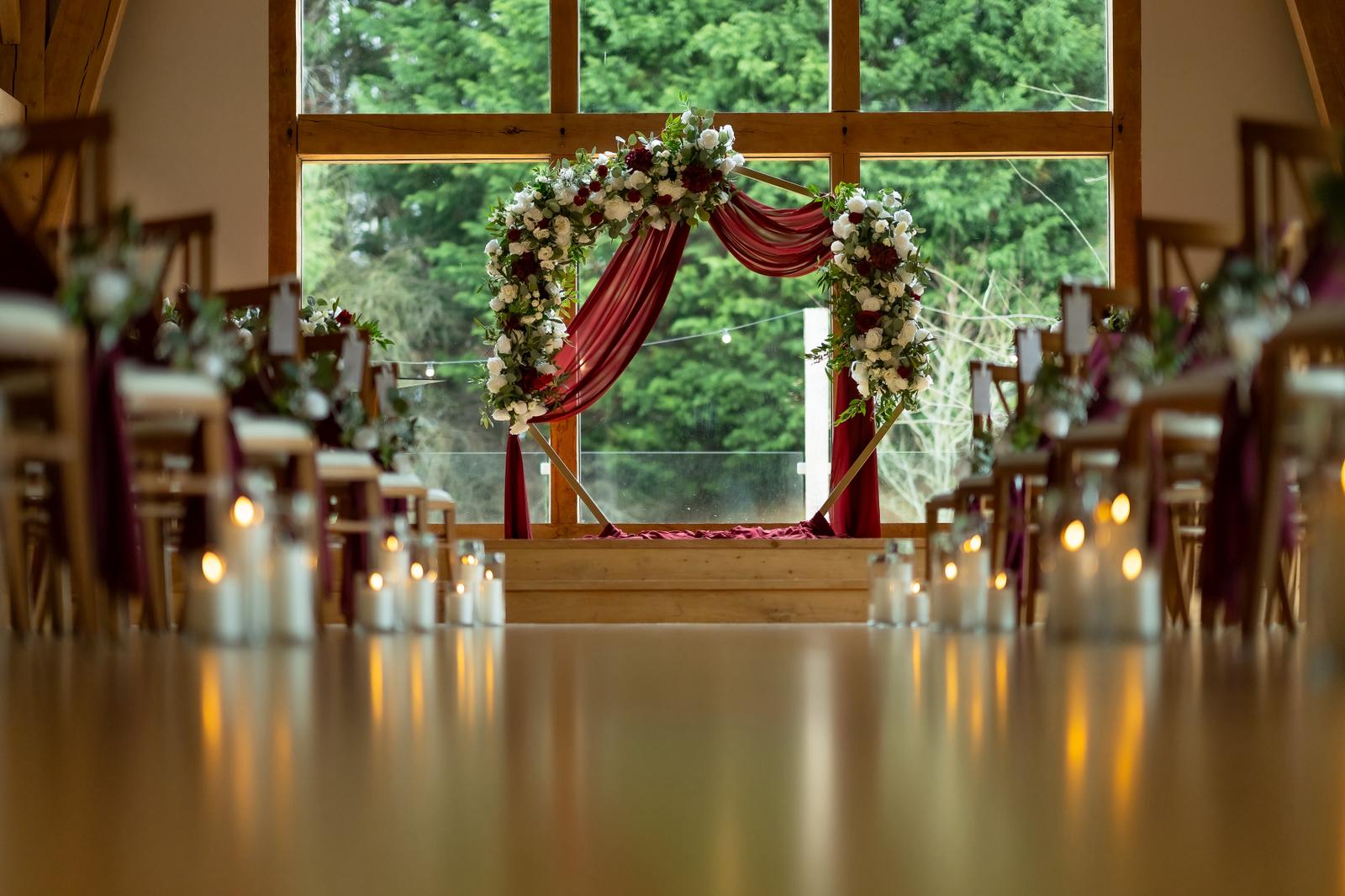Romantic burgundy wedding ceremony arch with floral draping and candlelit aisle at The Mill Barns