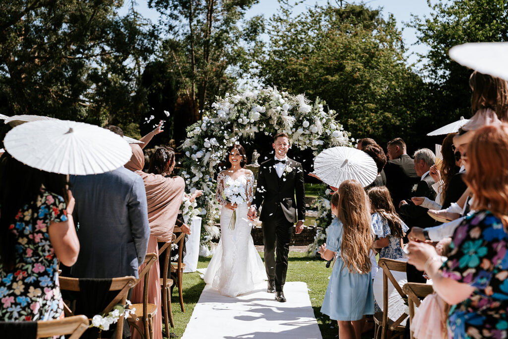 Outdoor wedding ceremony at Hogarths Stone Manor with bride and groom Humaira and David walking down the aisle beneath a white floral wedding arch with artificial wedding flowers. Wedding aisle decor Worcestershire.
