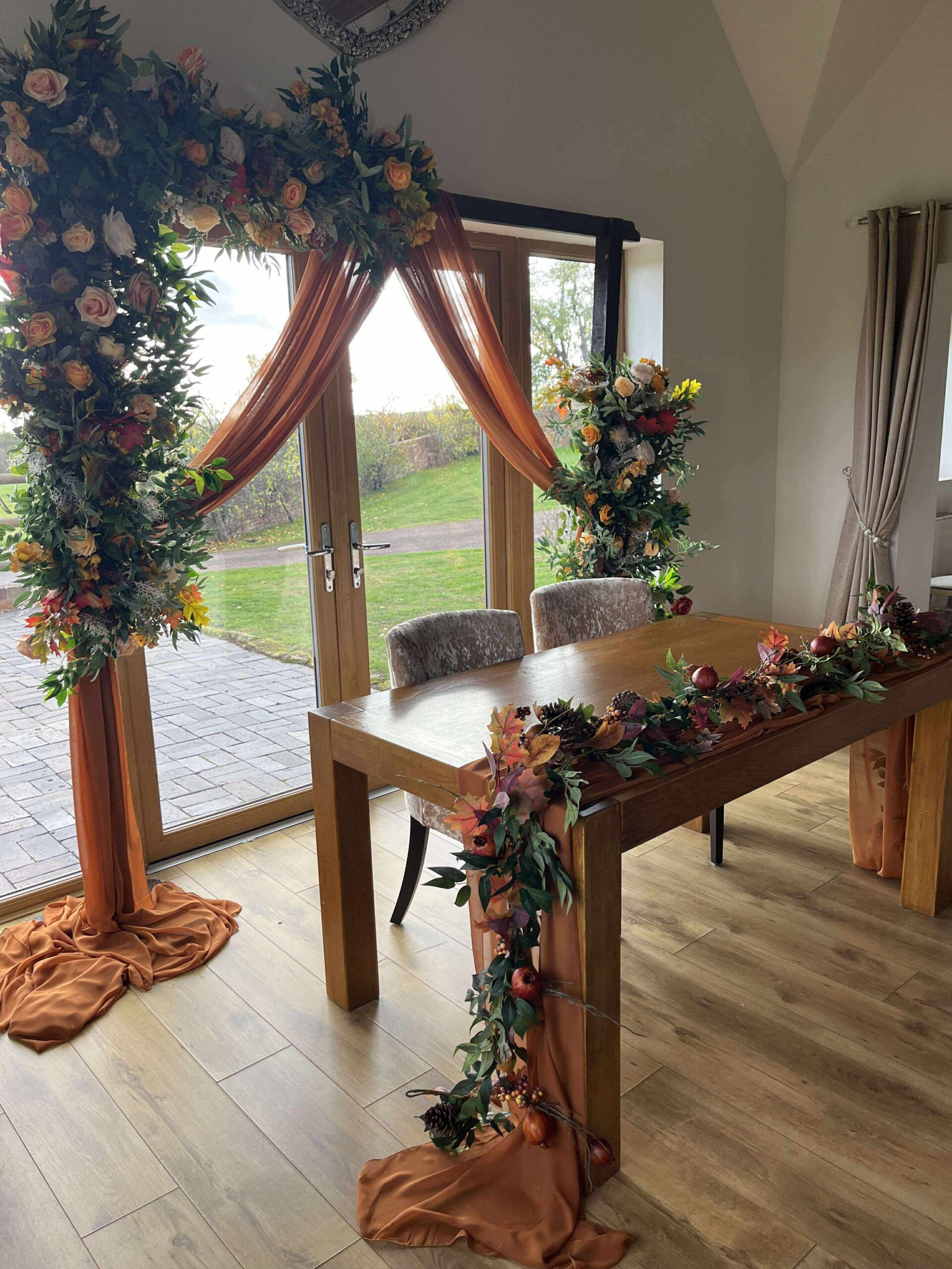 Rust draped wedding arch with autumn floral arrangement and foliage runner on wooden ceremony table at Bordesley Park