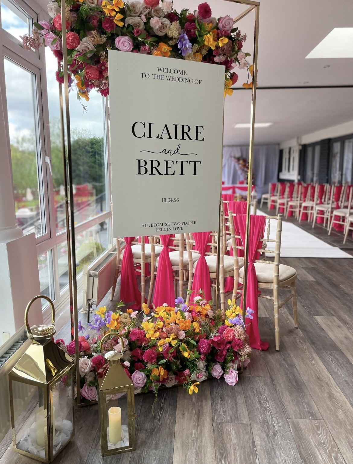Bright pink and orange wedding welcome sign with floral arrangement and gold stand at The Holt Fleet